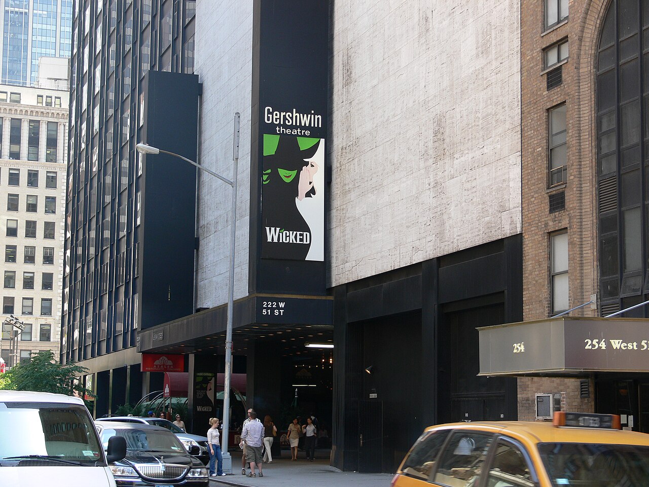 Street corner outside the Gershwin Theatre in New York City with a tall theatre banner and a yellow taxi