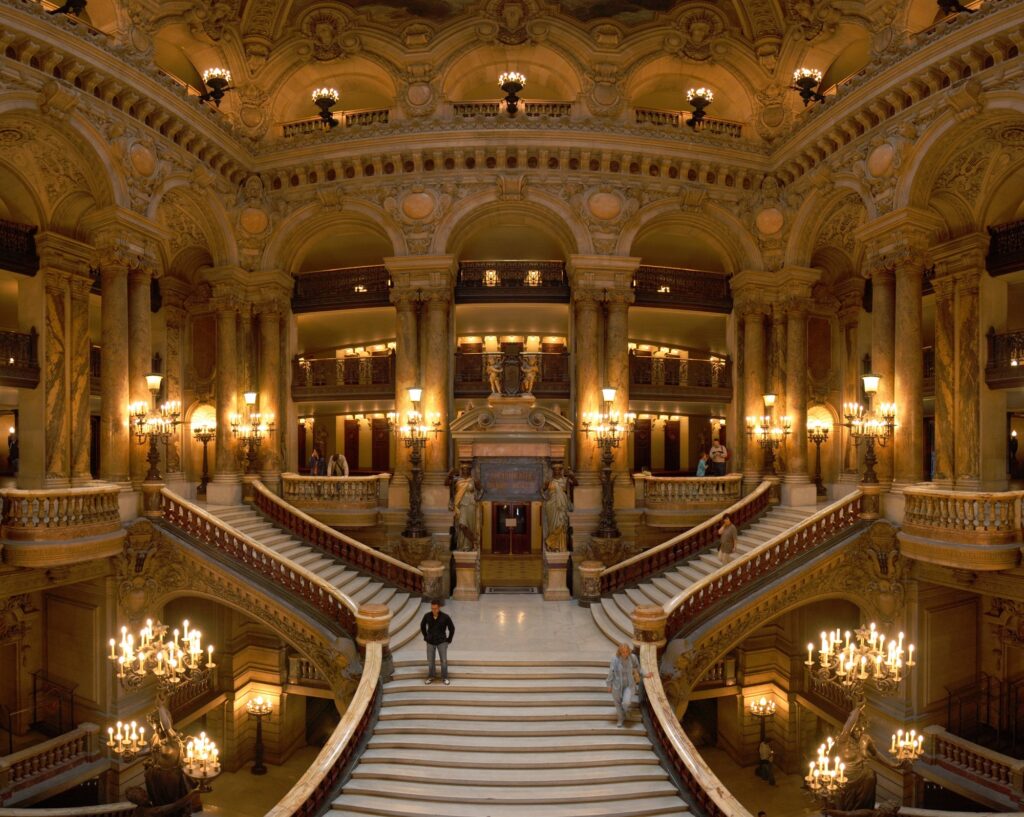 The marble Grand Staircase of the Palais Garnier opera house in Paris, lit by ornate candelabra beneath gilded arches