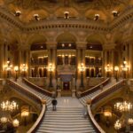 The marble Grand Staircase of the Palais Garnier opera house in Paris, lit by ornate candelabra beneath gilded arches