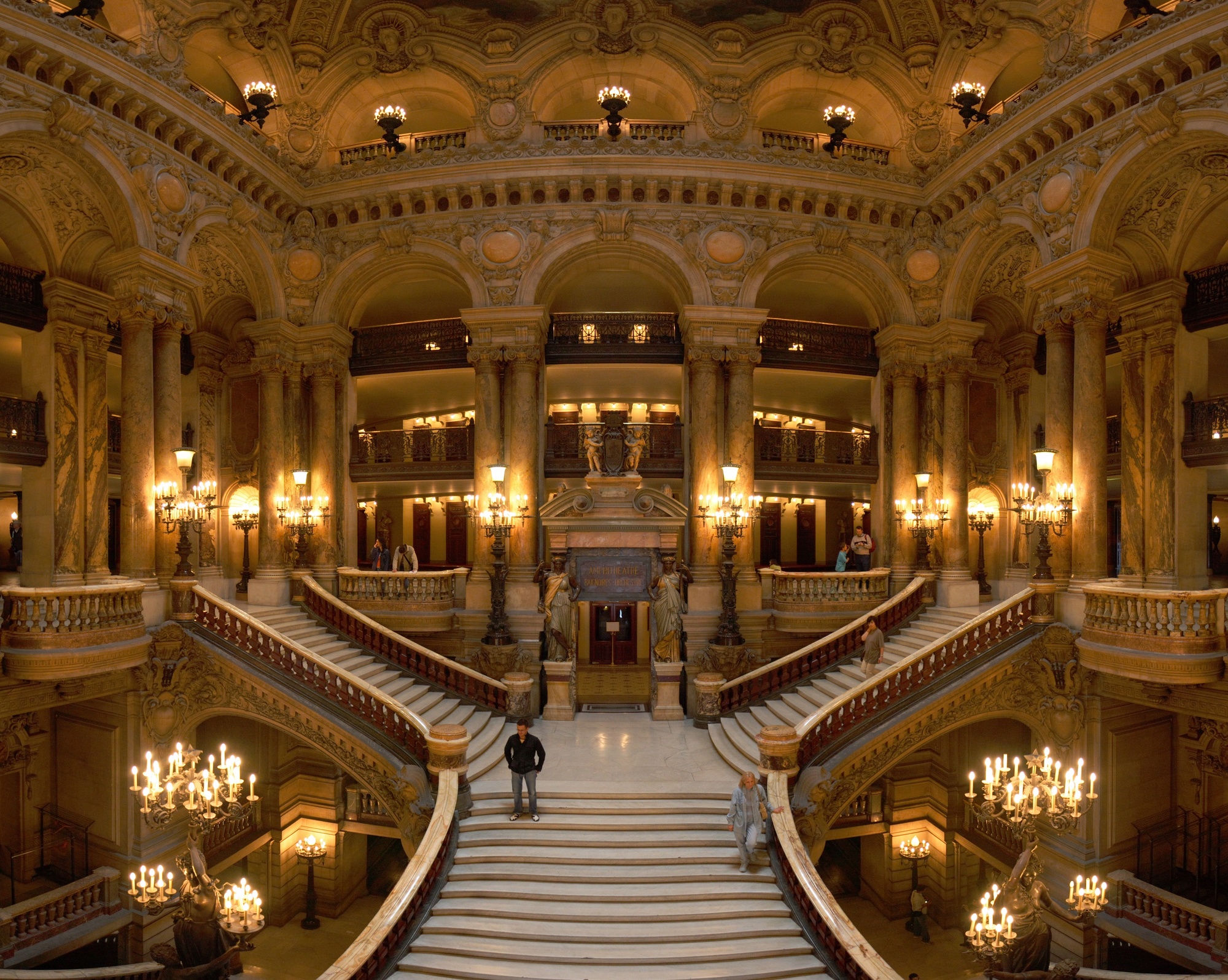 The marble Grand Staircase of the Palais Garnier opera house in Paris, lit by ornate candelabra beneath gilded arches